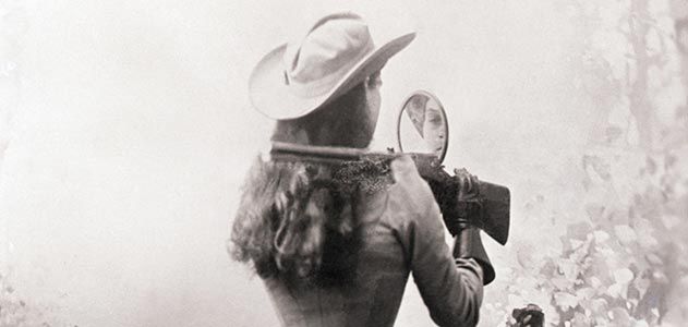 Rear view of woman looking into a mirror attached to a rifle that's resting over her shoulder