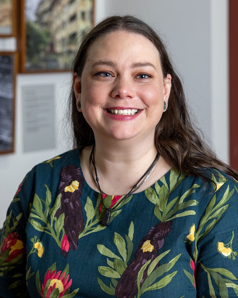 Head shoulders portrait of Jo, a white woman with long dark hair, smiling for the camera. She is wearing a blue shirt with black cockatoos and green leaves on it.