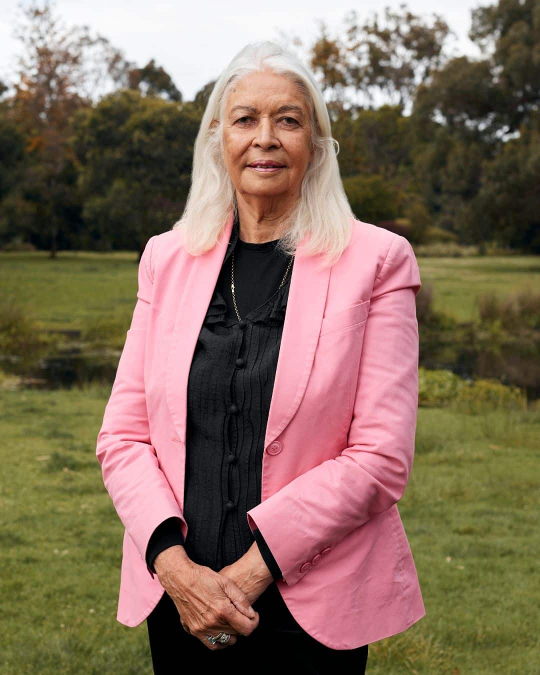 Photo potrait of Professor Marcia Langton AO. She is standing in a garden, surrounded by grass and trees. She wears a pink bazer and looks at the camera.