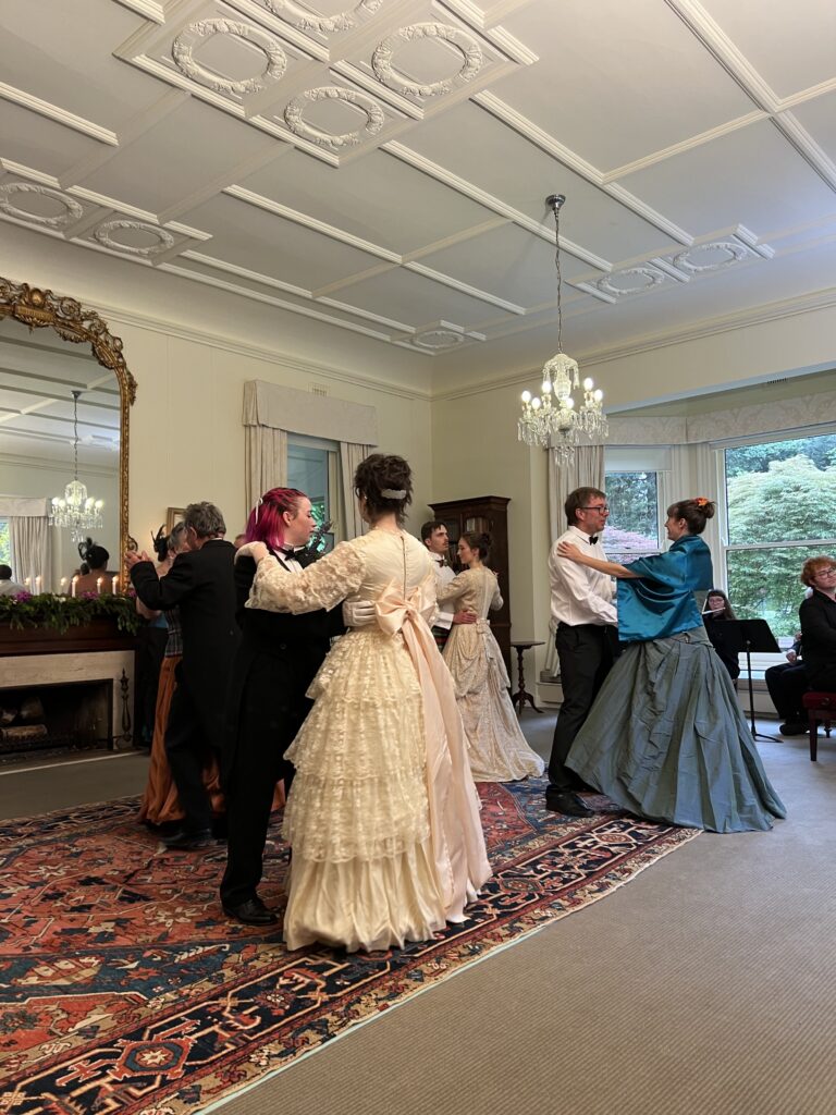 People dancing in pairs in what looks to be a fine mansion. Several of the women are wearing extravagent dresses with large skirts.