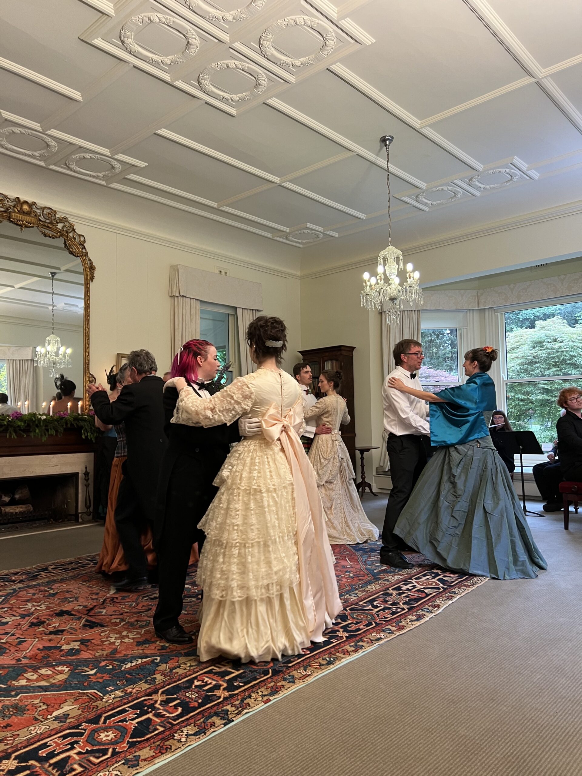People dancing in pairs in what looks to be a fine mansion. Several of the women are wearing extravagent dresses with large skirts.