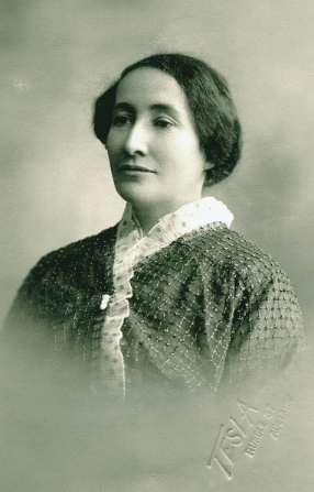 Head and shoulders studio portrait of a Grace Nell, sitting in three-quarter profile. Her hair is dark and her face placid as she looks off into the middle-distance.